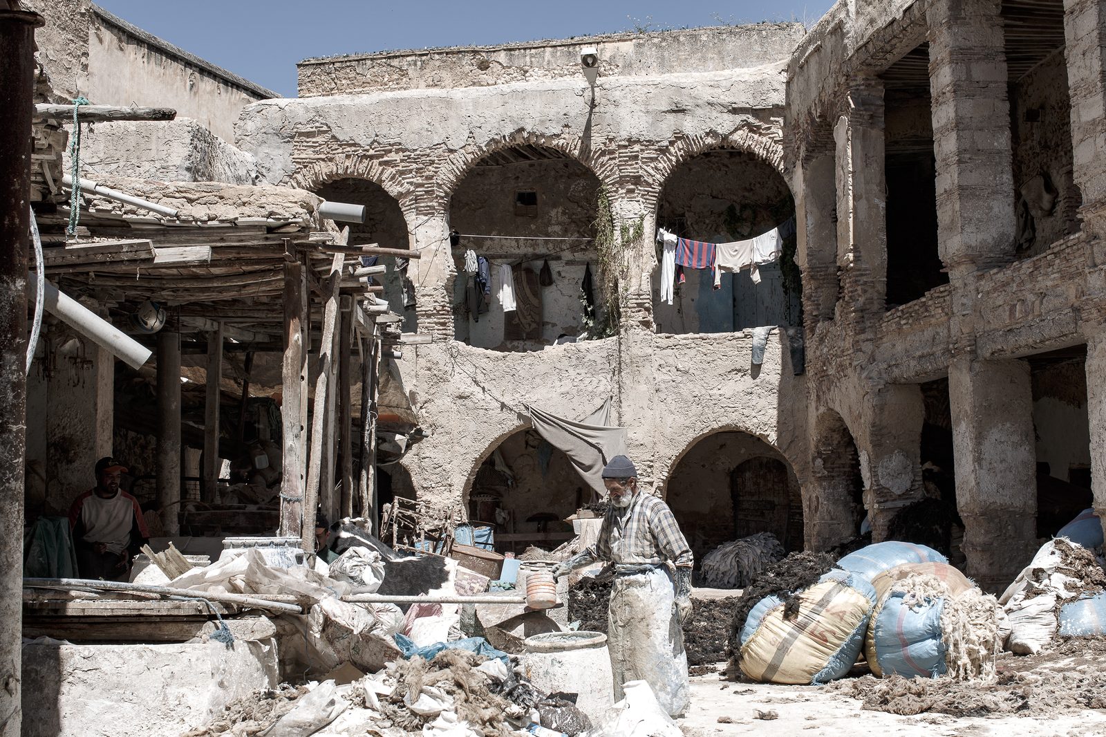 An elderly worker in a plaid shirt and stained apron stands in the sun-drenched courtyard of the Tanneries of Fès, surrounded by sacks of raw wool