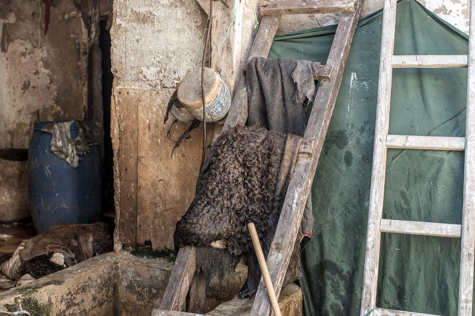 Animal hides drying on a wooden rack at an outdoor tannery, with a blue barrel and peeling plaster walls