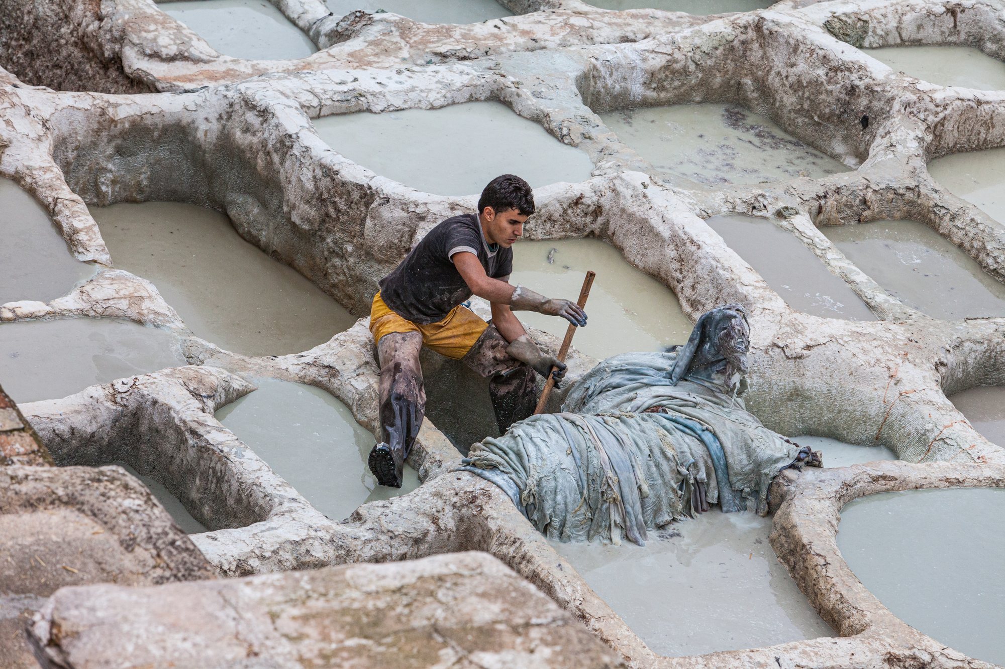 A tanner kneels at the edge of a vat, boots caked in tannery fluid