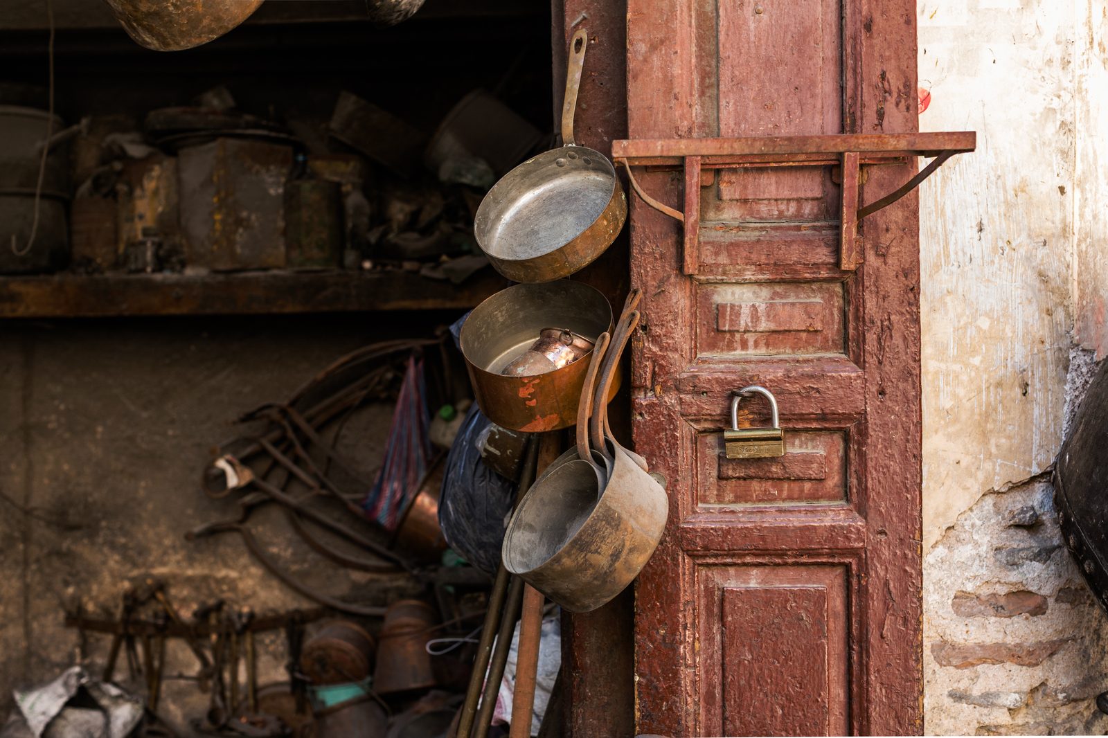 Hand-hammered copper cookware hanging on a weathered red wooden door frame in a Fès coppersmith workshop, a cluttered shelf of tools and a peeling wall behind
