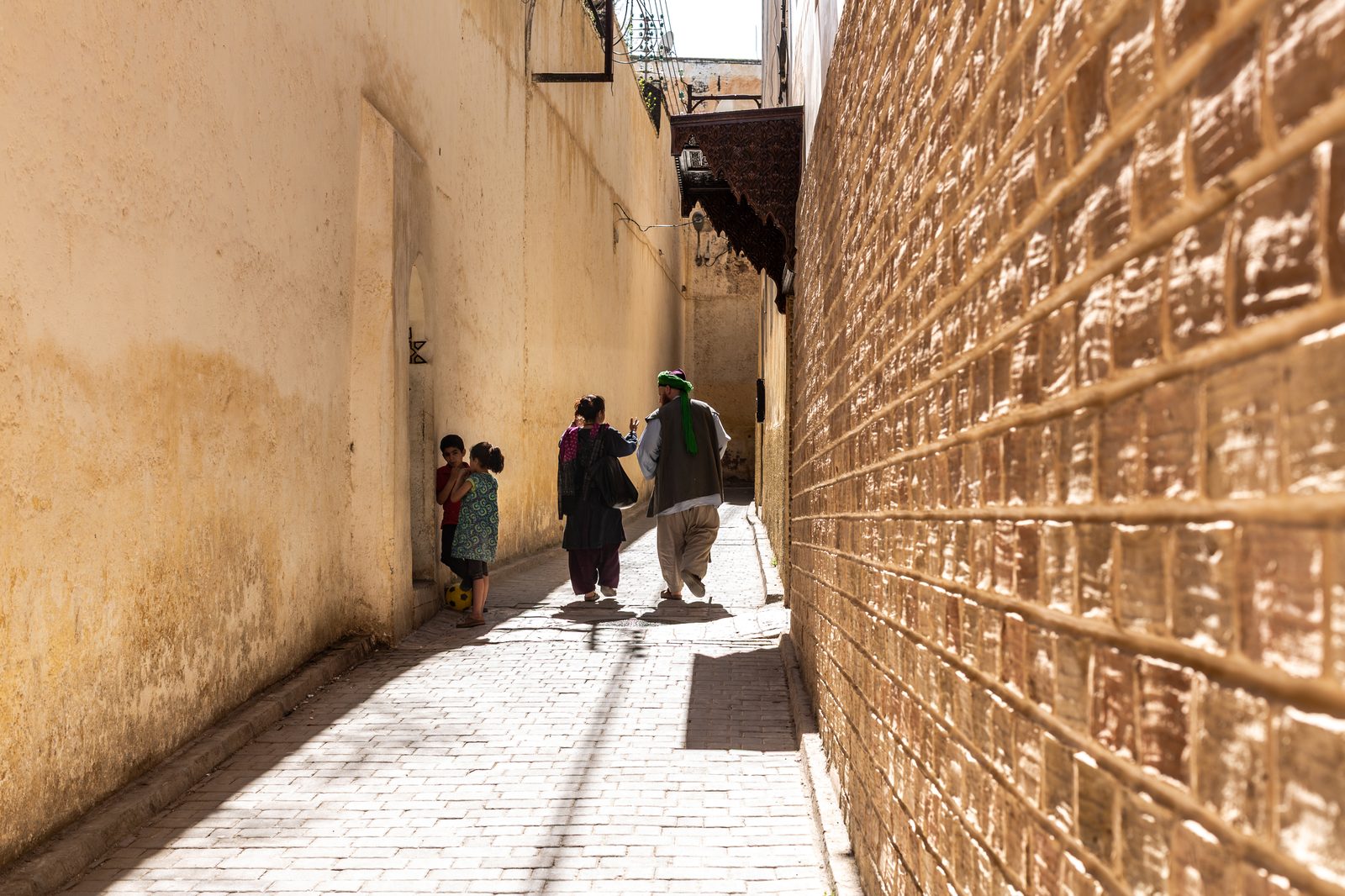 A sun-drenched narrow alley in the Fès medina, a man in a green fez and a woman in a dark coat walking away, two children by a doorway with a soccer ball at their feet and a carved wooden balcony above