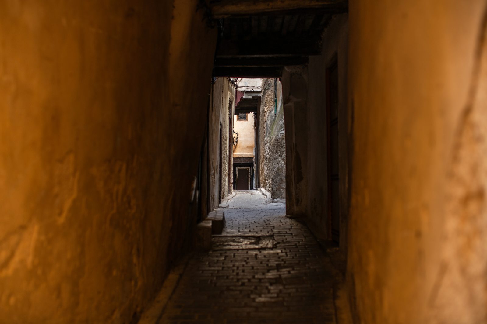 A view down a narrow covered derb in the Fès medina, textured sun-bleached earthen walls and a dark wooden overhead beam leading to a sunlit courtyard