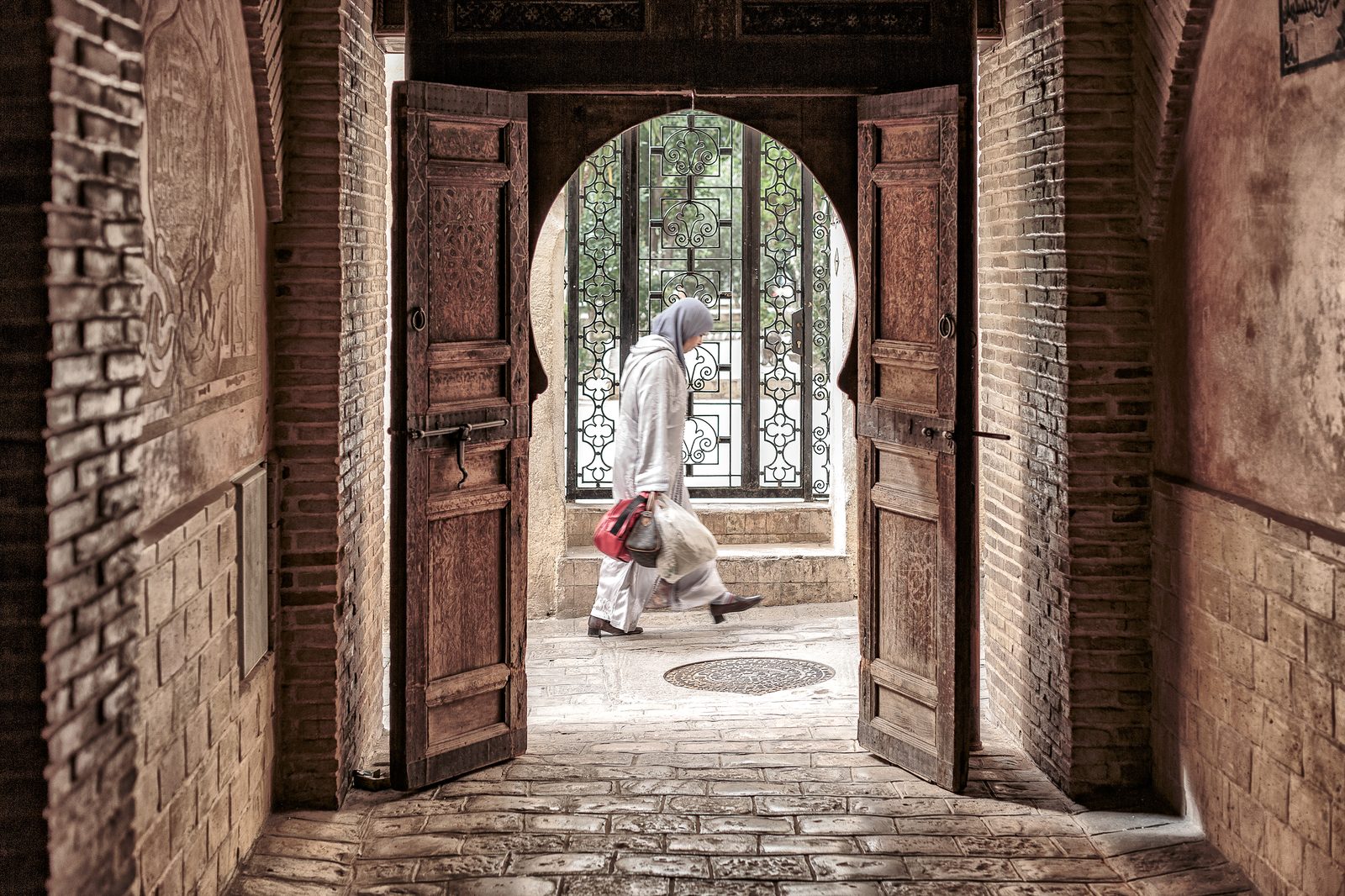 A woman in a white robe and headscarf walking through a grand carved wooden doorway, framed by brick walls and trees and an ornate iron gate