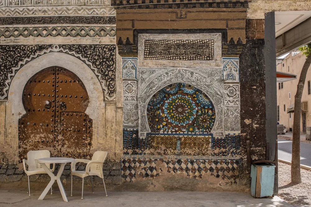 A weathered facade in the Fès medina: Moorish stucco carvings, a studded wooden door, and a zellige mosaic arch beside modern patio chairs