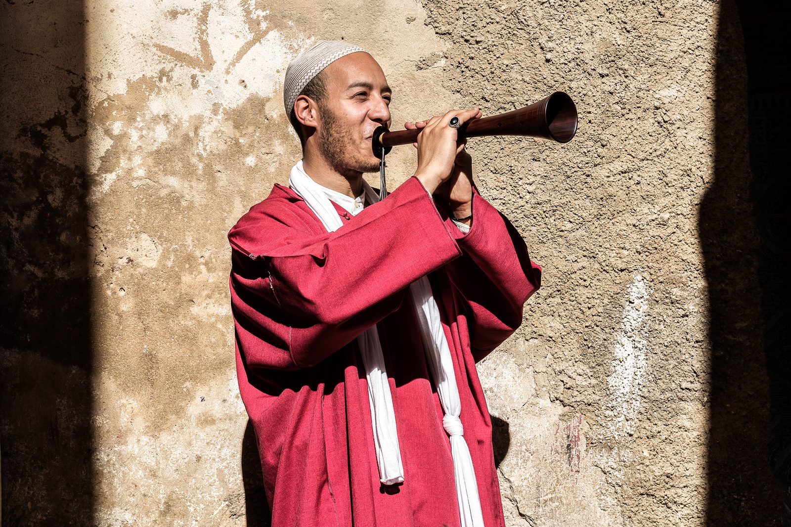 A musician in red robes and a kufi cap playing a zurna, smiling, against a sun-drenched textured plaster wall