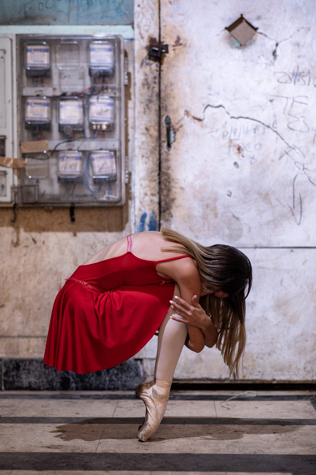 A ballerina in a red dress in a deep plié against a weathered wall and an electrical meter box