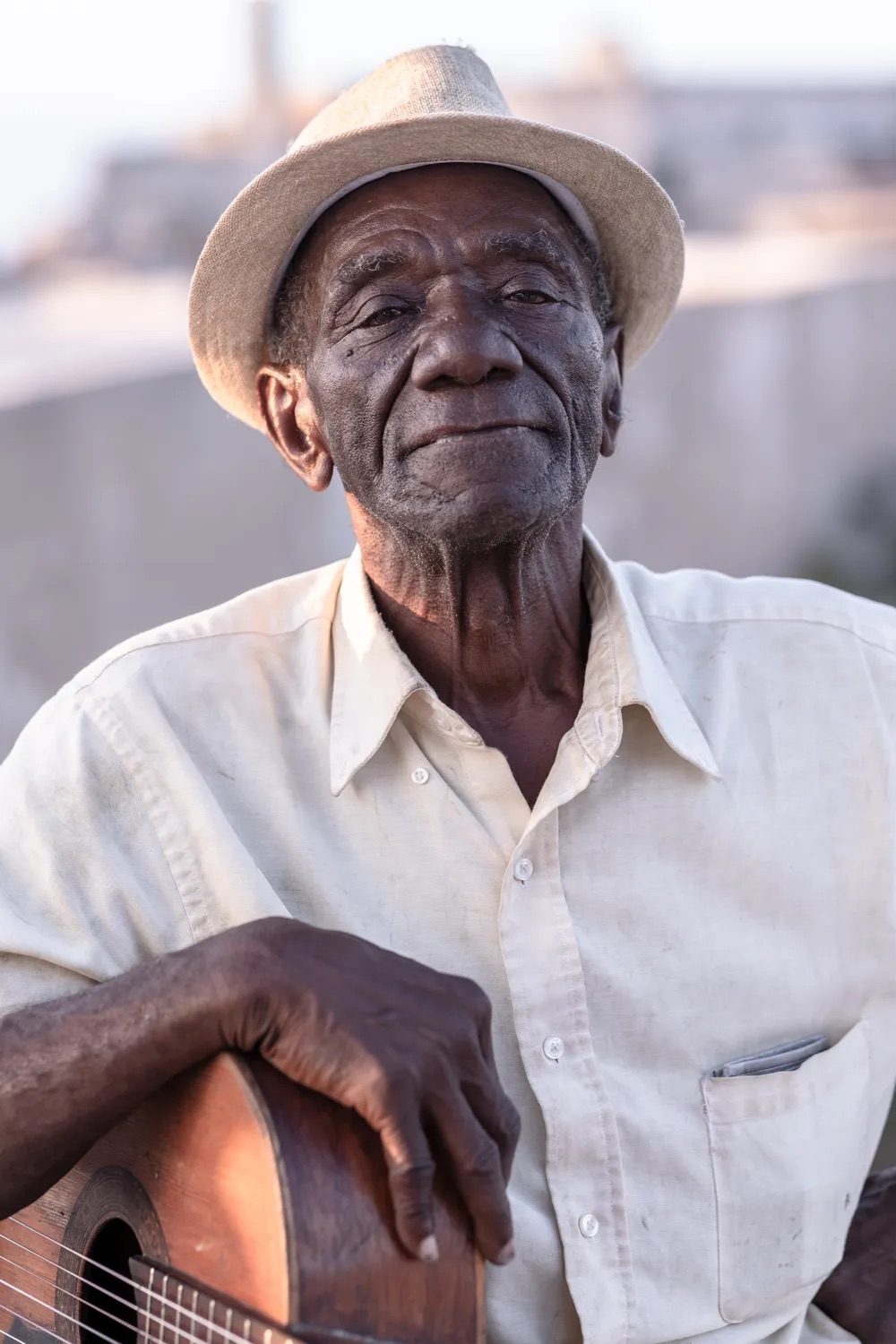 An elderly street musician in a straw hat resting his hands on a wooden guitar