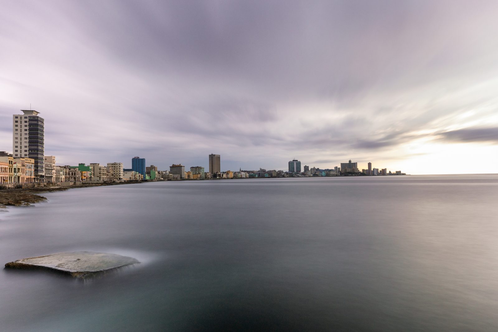 A long-exposure panorama of the Havana skyline along the Malecón, colonial and mid-century towers above silky calm water under a streaky sky
