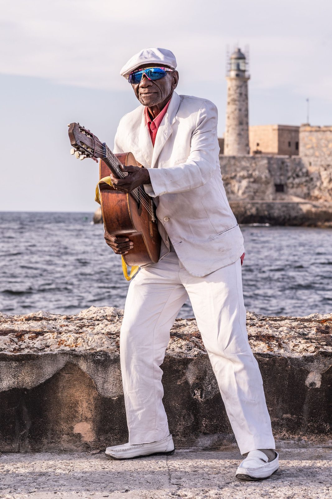 A smiling man in a white suit playing guitar on a sea wall, the El Morro lighthouse behind him