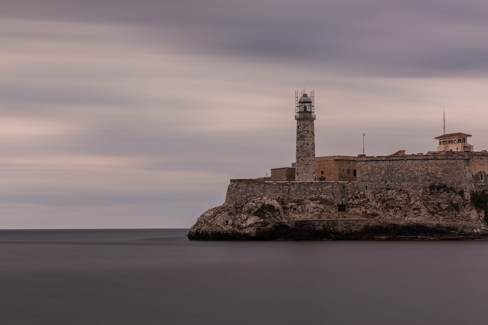 A long-exposure of the El Morro Castle lighthouse and fortress walls on a rocky outcrop, standing against a soft cloudy twilight sky