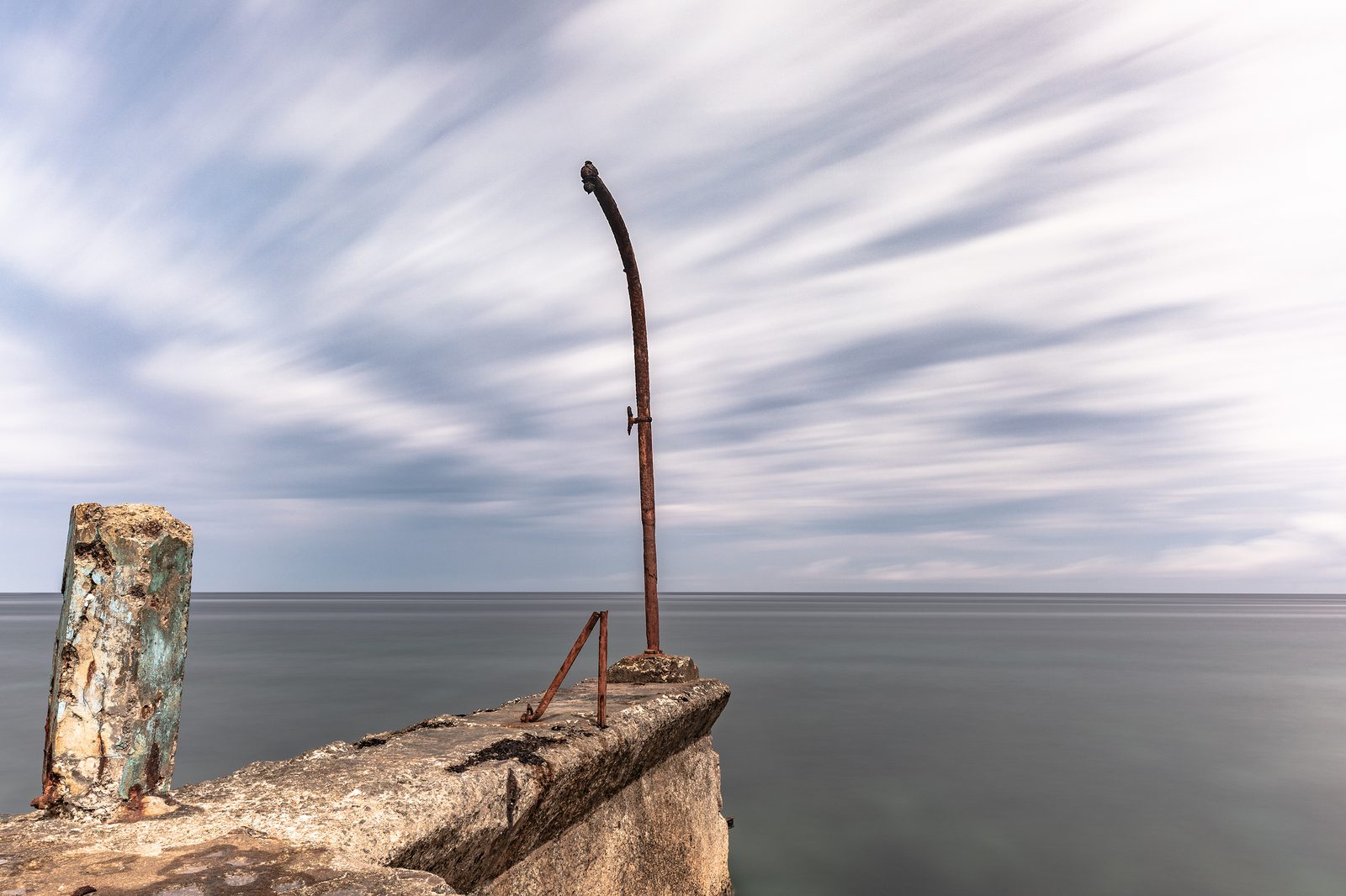 A weathered concrete pier and rusted metal post on the Malecón, overlooking a calm sea and streaky long-exposure sky