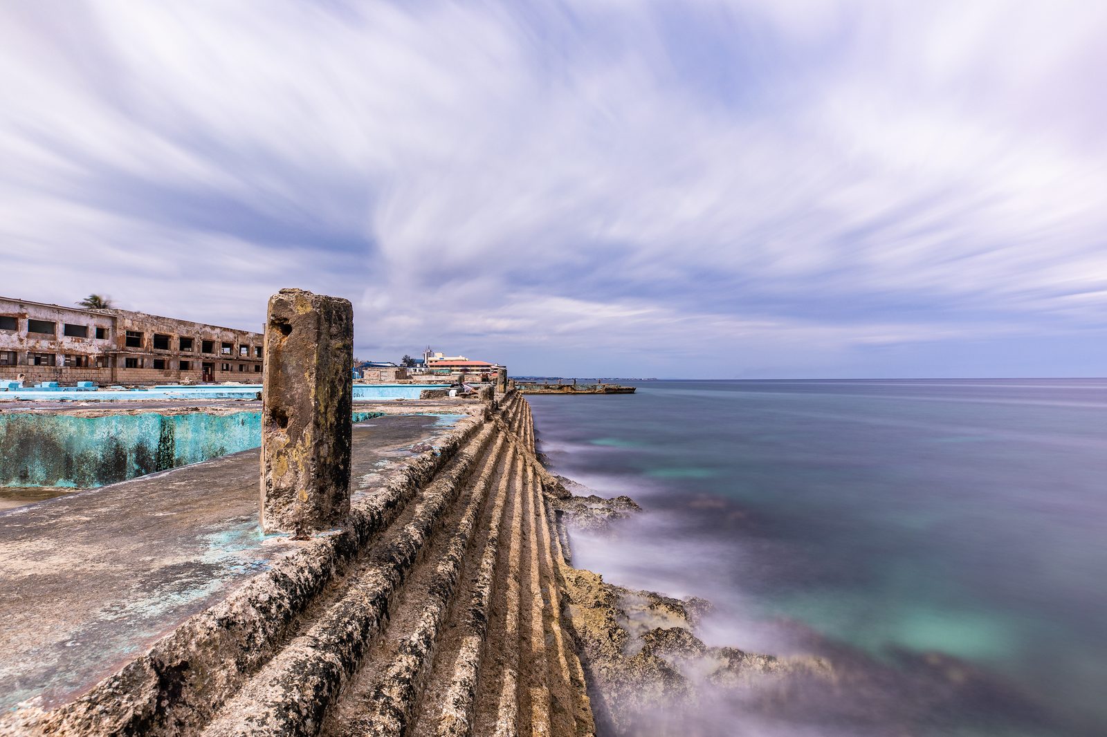 A derelict mid-century building with a turquoise pool beside a weathered sea wall, the silky ocean meeting a streaky sky