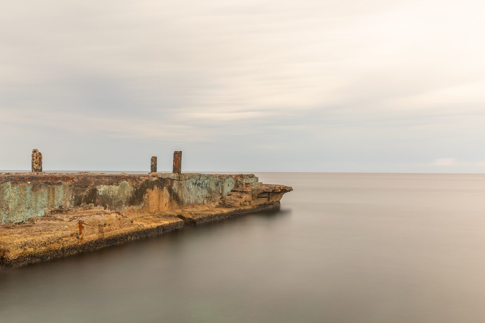 A weathered concrete pier extending into a calm grey sea under an overcast sky, the textured decay of Havana's coastline