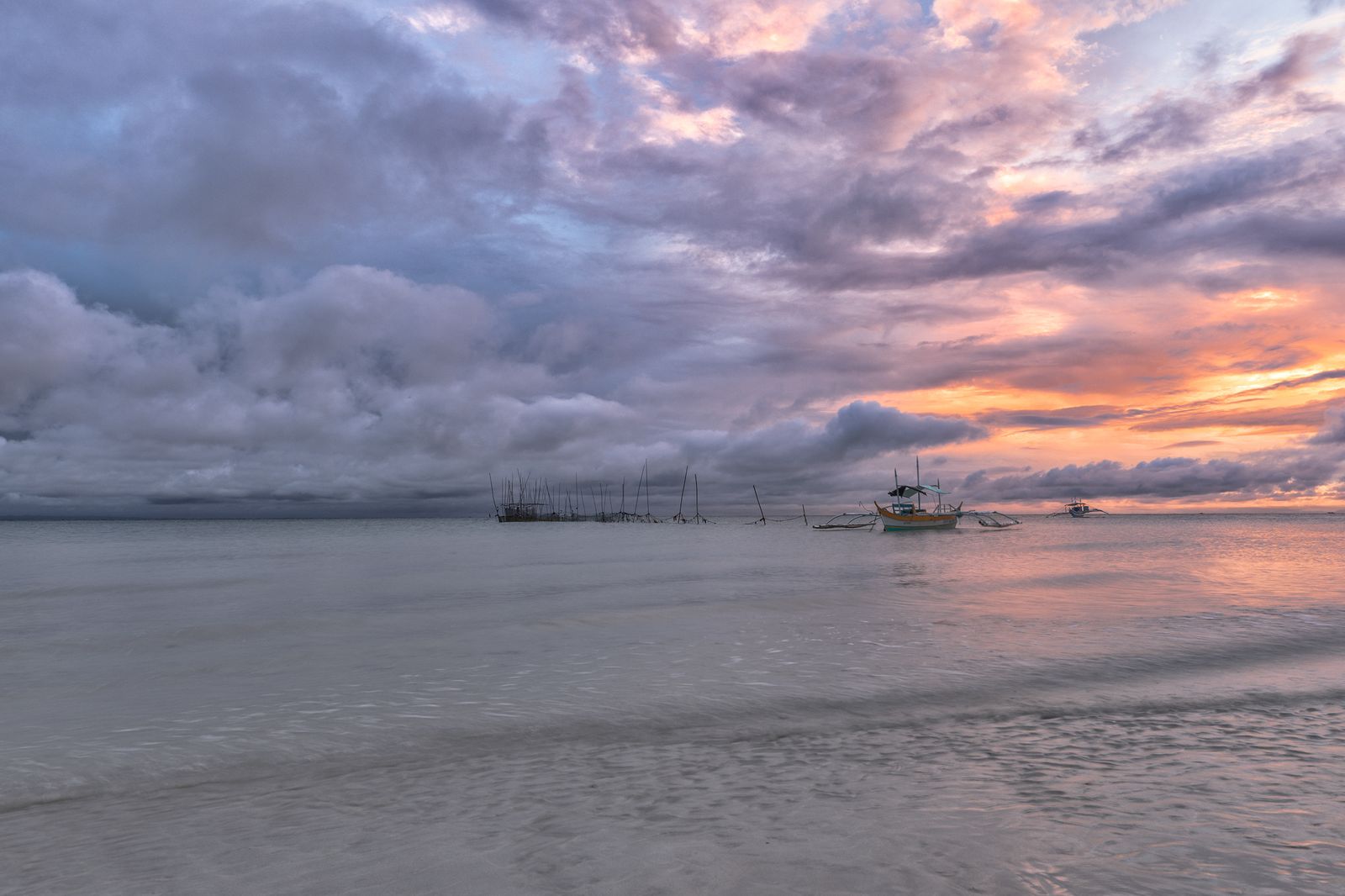 A traditional outrigger boat and fish traps at twilight against a dramatic, cloud-filled sky painted with sunset hues.