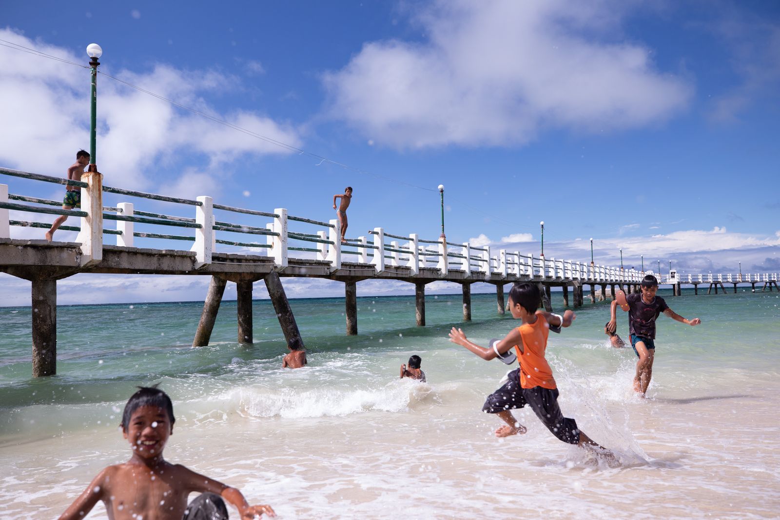 Children playing in the turquoise surf beneath a long, white-railed pier on a sunny tropical day.