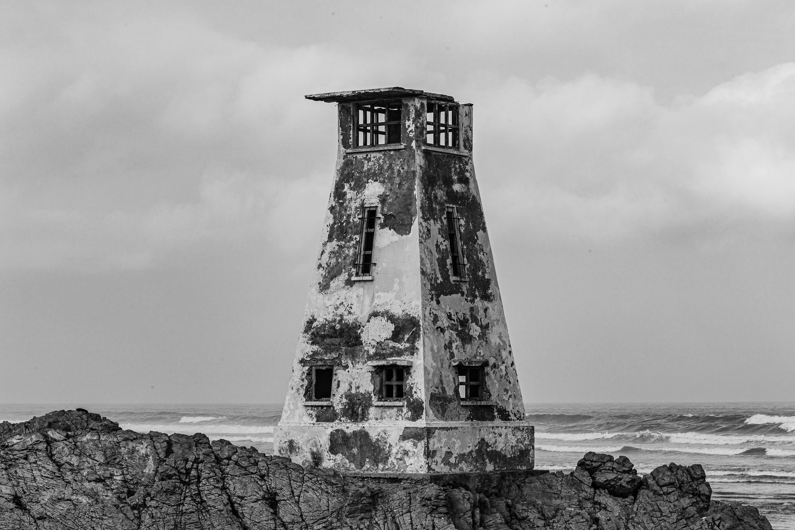 Monolith I - a weathered square lighthouse tower on a jagged rocky outcrop, its peeling facade against an overcast sea