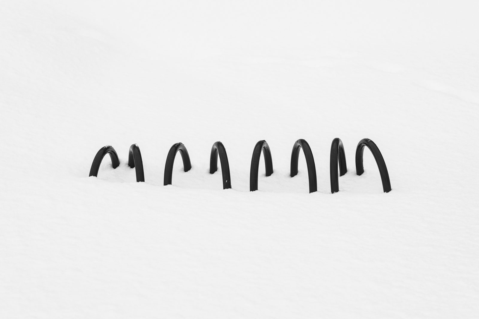 A row of black bicycle racks protruding from deep snow, a stark horizontal line on white