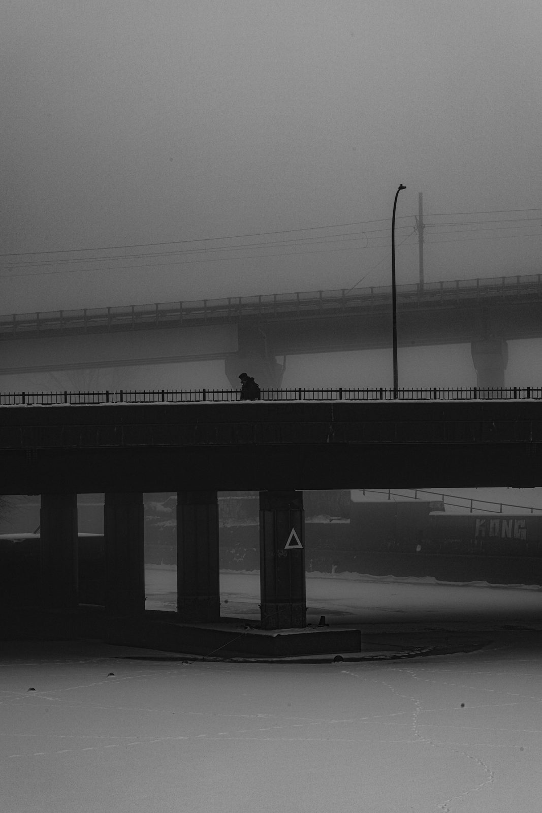 A solitary figure walking a concrete bridge in fog, framed by industrial structures and snow