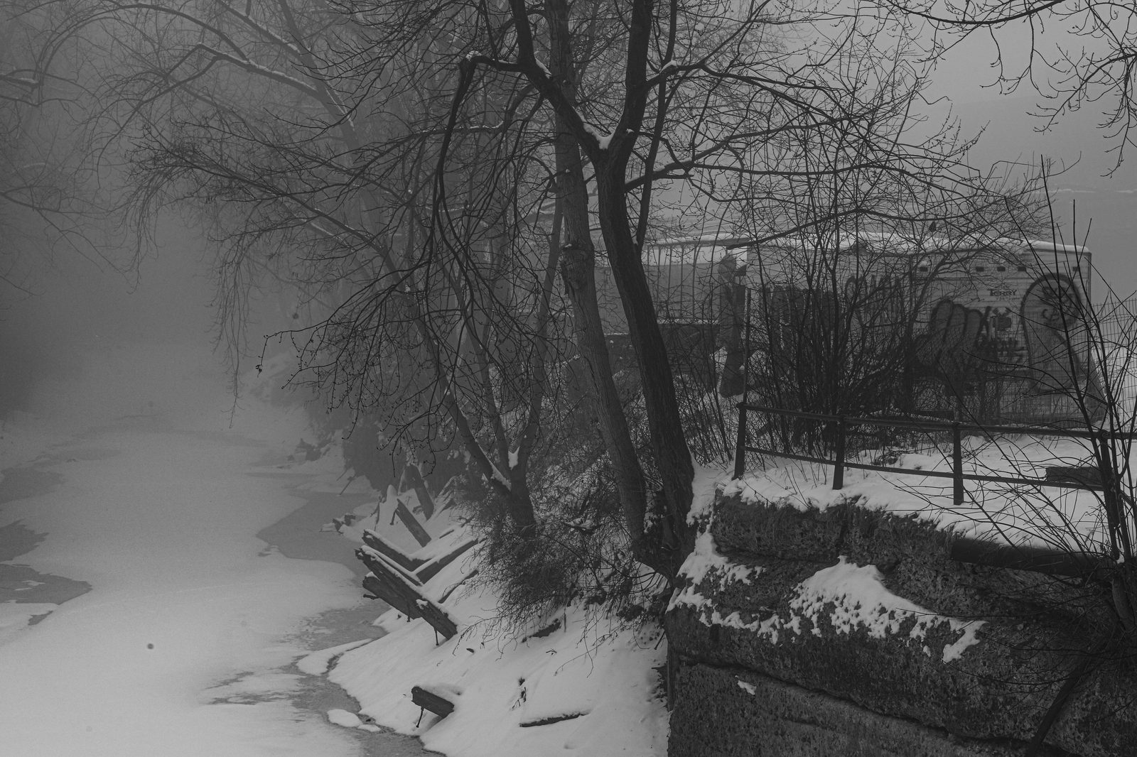 A frozen canal bank framed by bare trees, a snow-dusted stone retaining wall and railing lining icy water, a graffiti-covered building faint through the fog