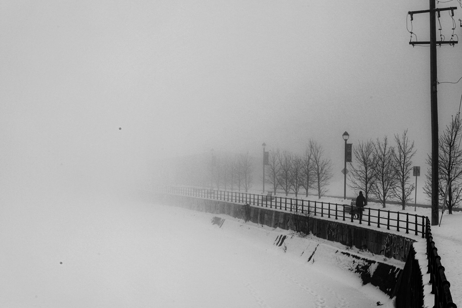 A figure walking a dog along a snow-covered canal path, fading into thick winter fog
