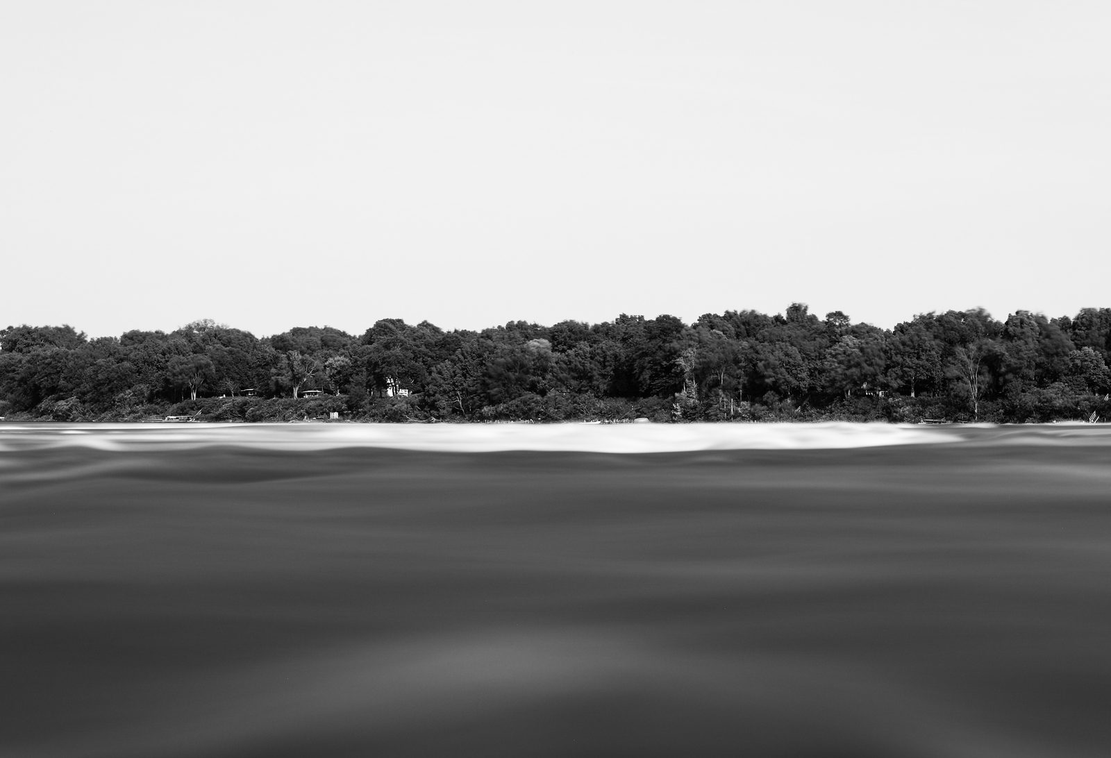 A long-exposure of a silky river below a dense dark forest shoreline and a featureless winter sky