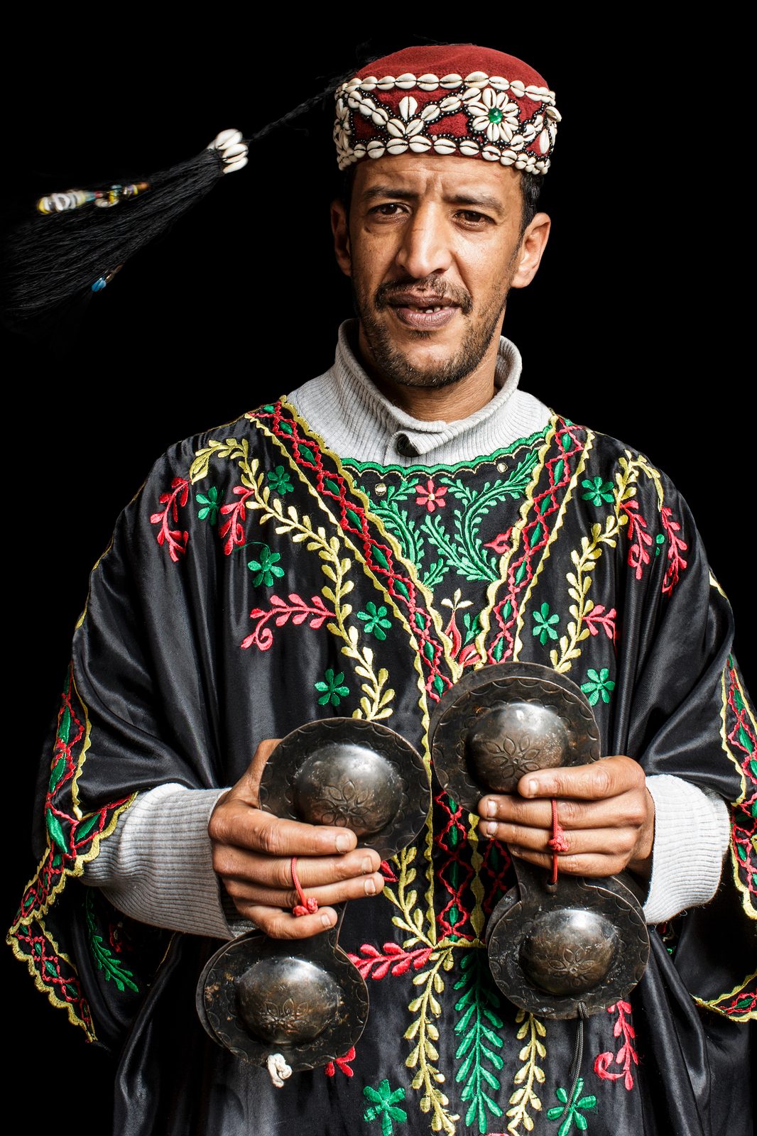 A Moroccan Gnawa musician in a red cowrie-shell hat and black embroidered robe, holding krakebs