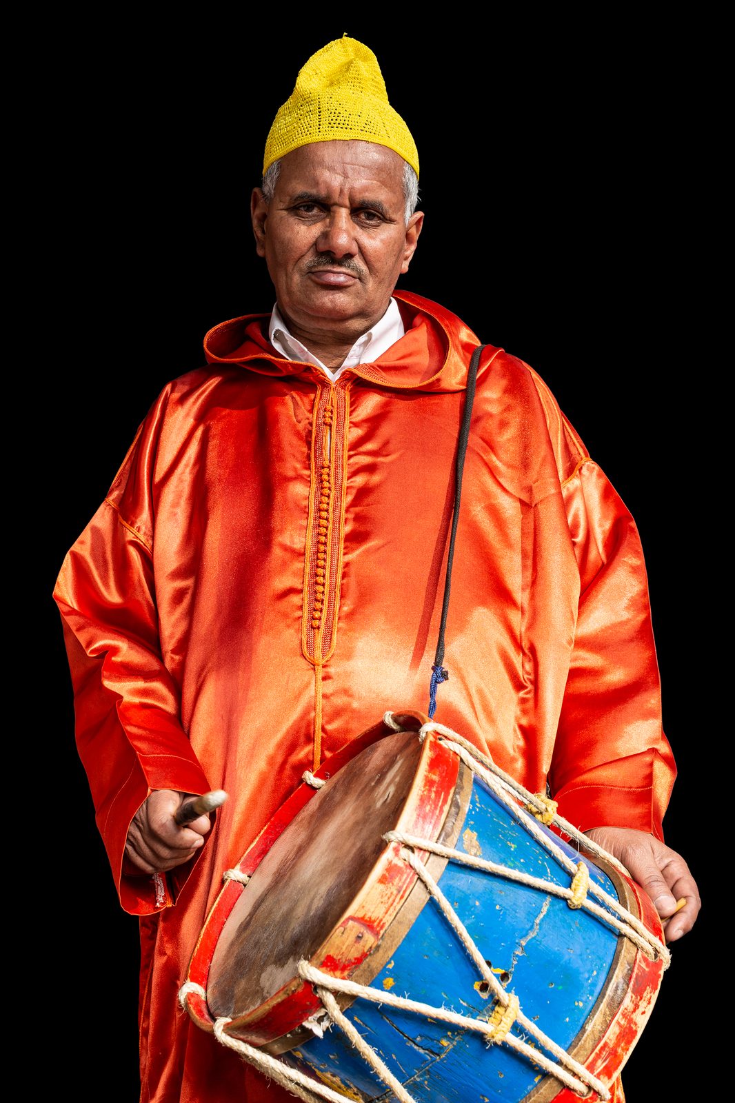 An elderly Moroccan man in a yellow cap and orange hooded robe, holding a blue and red drum