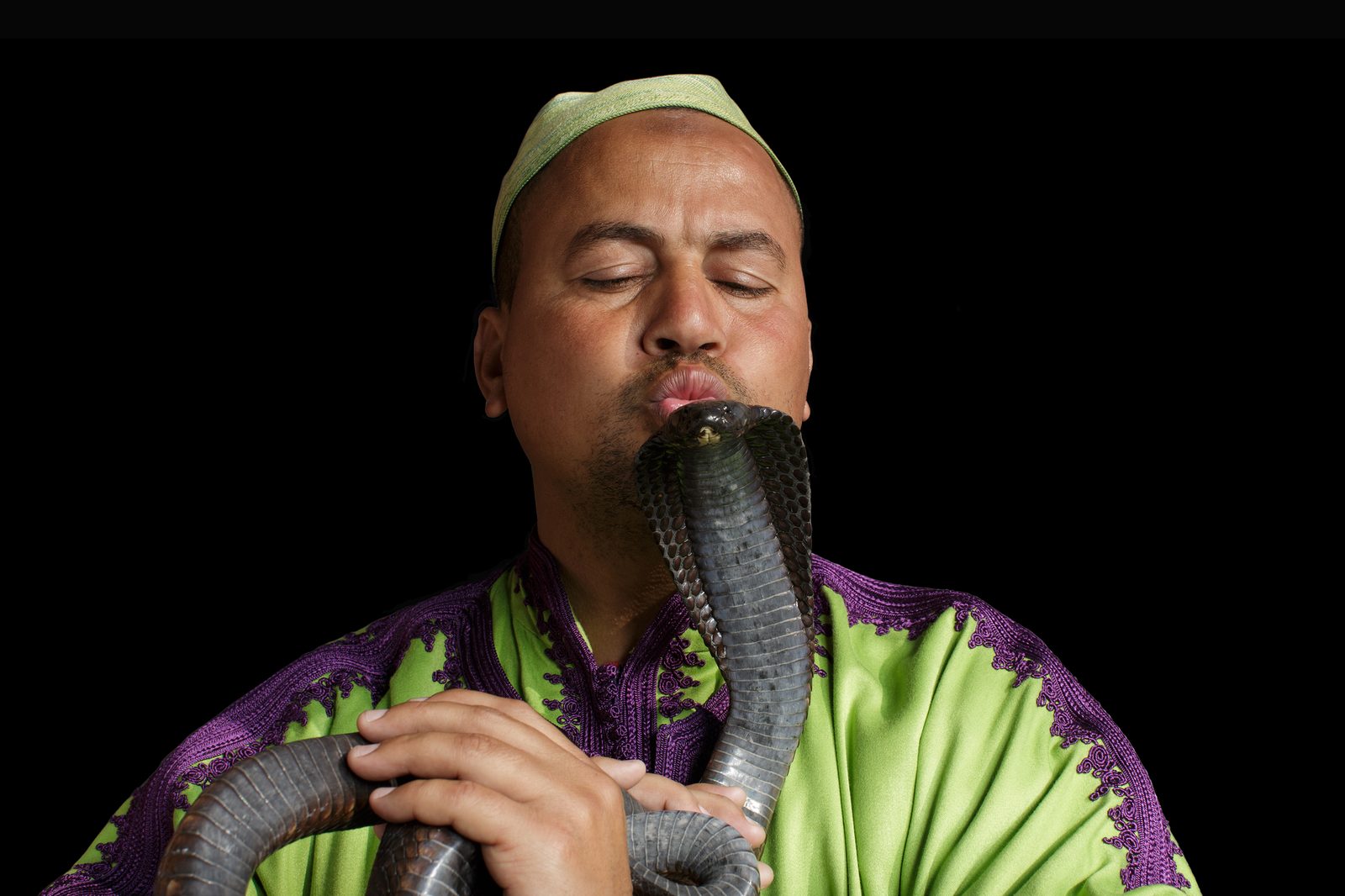 A Moroccan snake charmer in green and purple embroidered attire, a cobra raised to his closed eyes