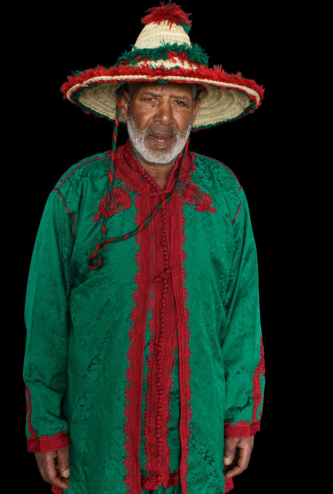 An elderly Moroccan Amazigh man in a wide-brimmed straw hat trimmed with red and green yarn and an emerald-green robe with red tarz embroidery, photographed against black