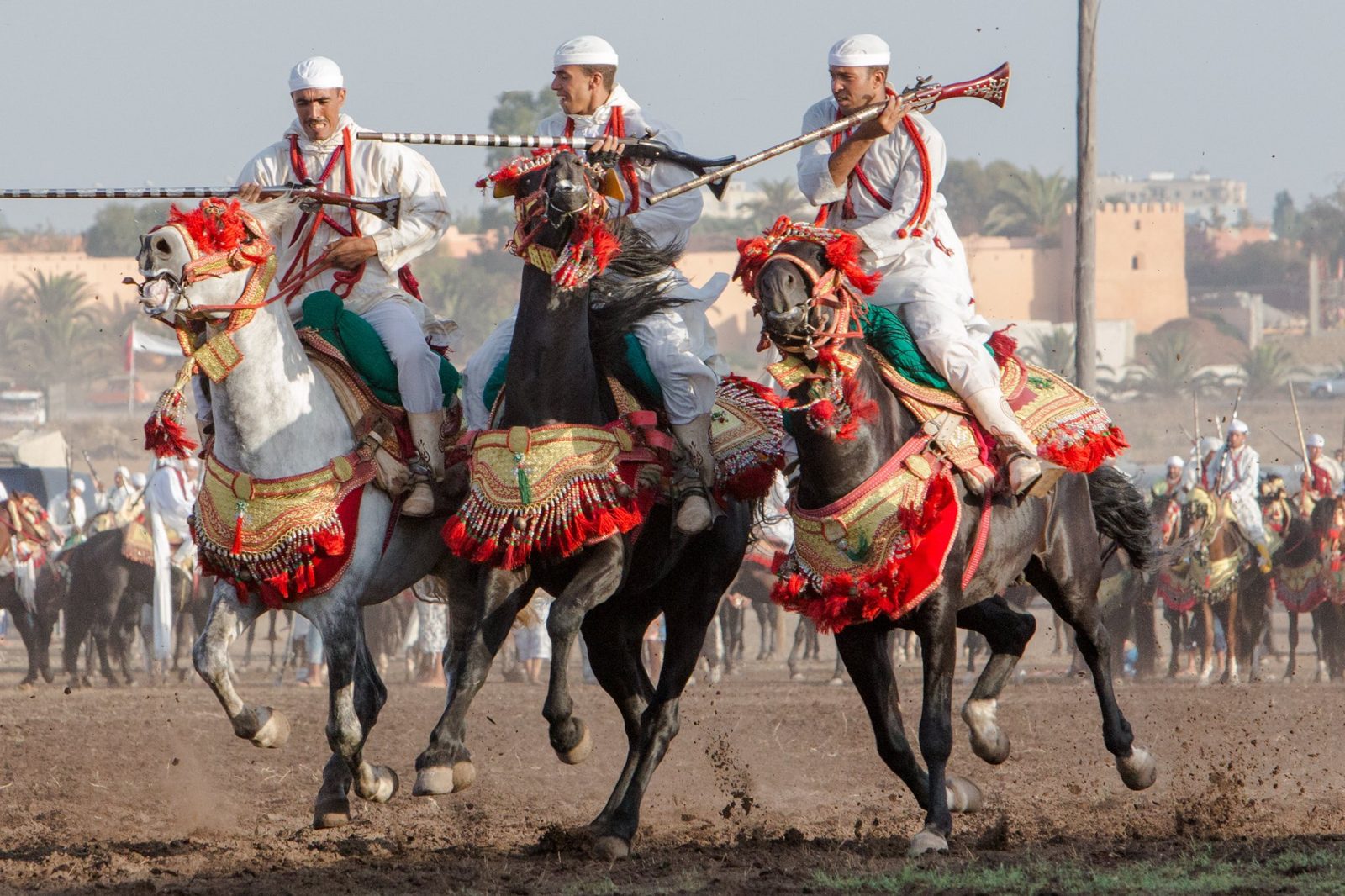 Three riders mid-charge, decorated horses kicking up earth, rifles held across the body