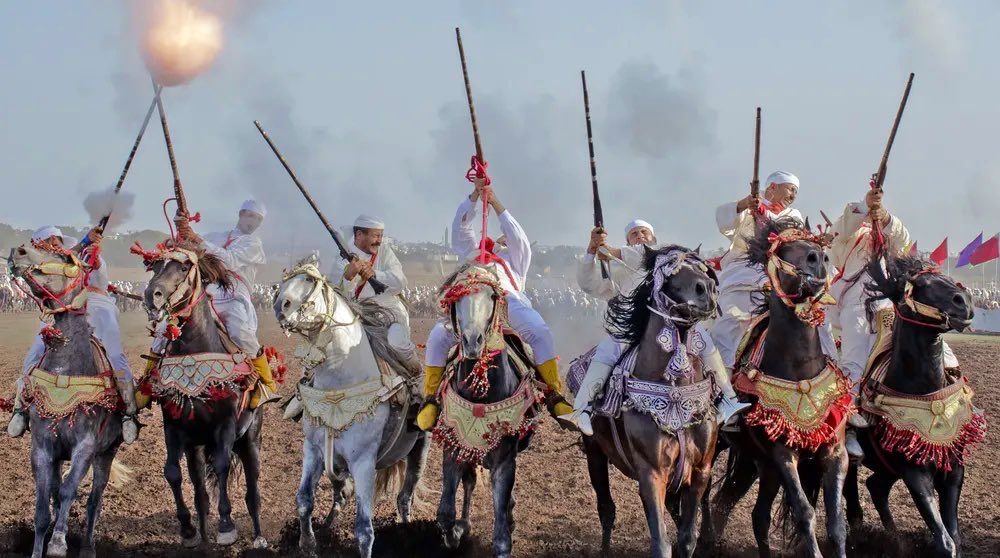 A line of riders in white firing in unison, smoke and muzzle flash at the front of the charge