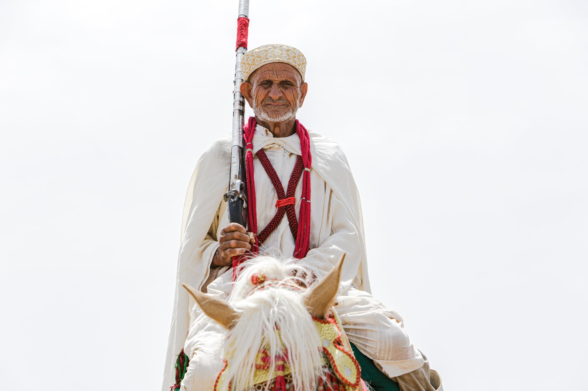 An elderly horseman in white, holding a moukhala, beside a white horse