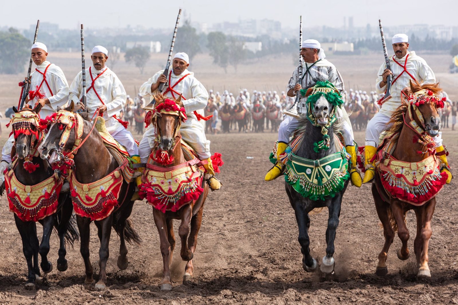 A formation of white-robed riders holding moukhalas aloft, kicking up dust in a coordinated canter